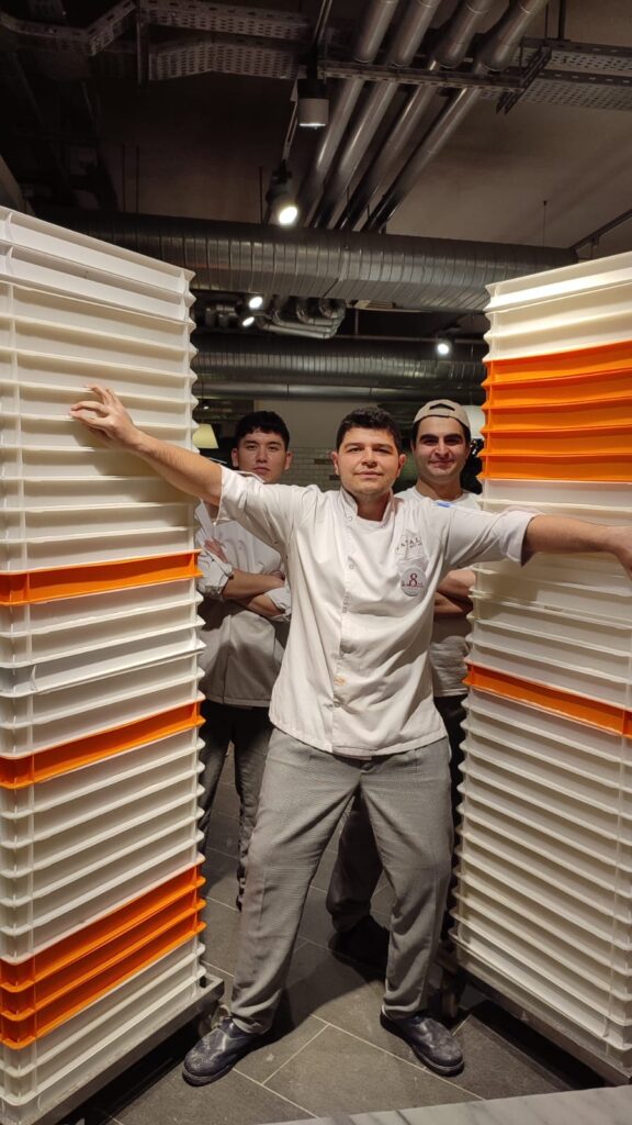 Professional chef Altaycan posing confidently with his kitchen crew between towers of fermenting dough trays for large-scale pizza production.
