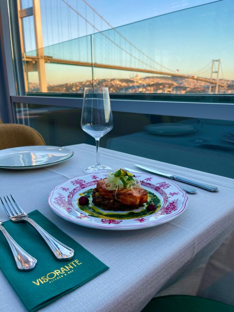 A gourmet seafood dish at Visorante Kitchen & Bar in Istanbul, featuring a perfectly seared salmon over a bed of lentils and greens, with a stunning view of the Bosphorus Bridge in the background.