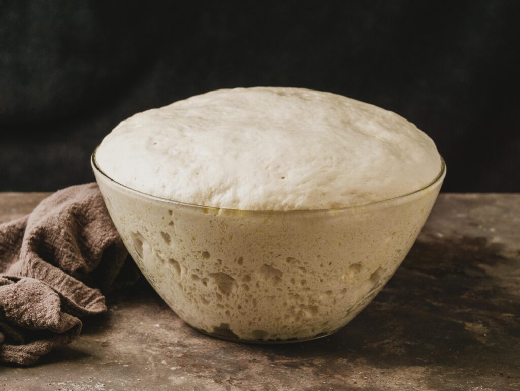 high angle bowl with growing dough pizza, A macro view of a high-hydration pizza dough undergoing a 48-hour cold fermentation process. The yeast has created tiny carbon dioxide bubbles, which are essential for achieving that light, airy, and open crumb structure known as the cornicione in a true Neapolitan pizza.