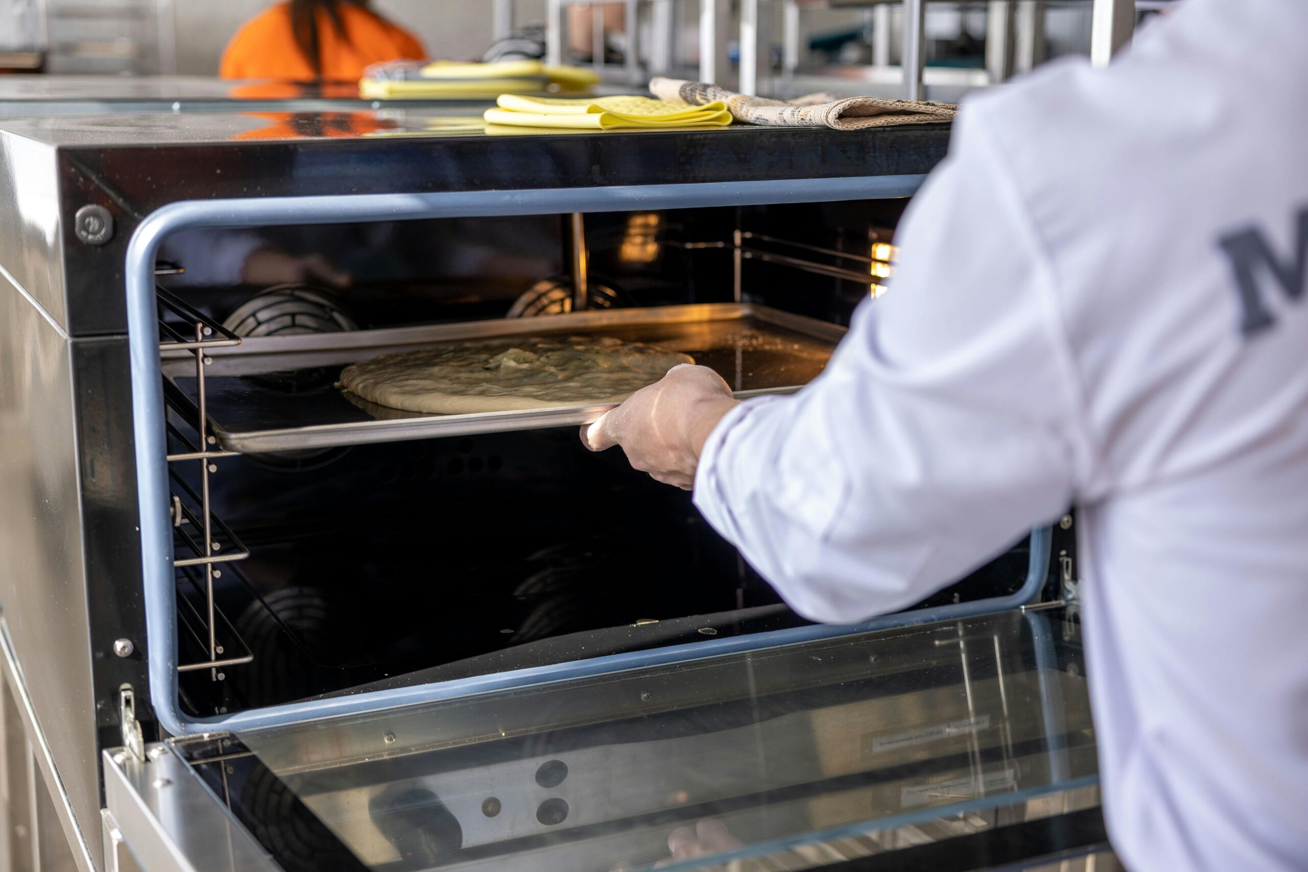 Chef places uncooked pizza into an industrial oven in a professional kitchen setting.