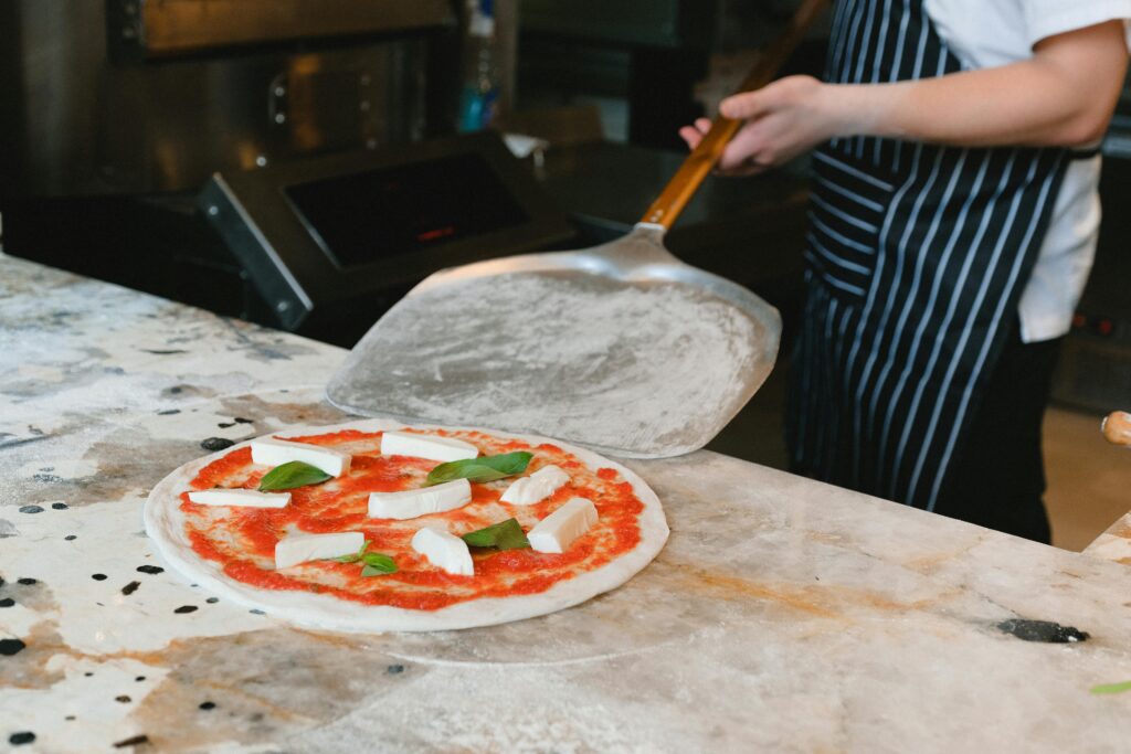A chef prepares uncooked pizza with cheese and basil on a marble counter.